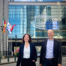 Carla Liehr und Tobias Möller-Walsdorf stehen vor dem Europäischen Parlament in Brüssel. Beide lächeln in die Kamera. Im Hintergrund wehen die Flaggen verschiedener europäischer Staaten.