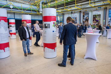 Das Bild zeigt eine große Halle. Im Vordergrund stehen mehrere Menschen um eine Litfaßsäule herum, auf der Text und Bilder abgedruckt sind. Der Hintergrund zeigt eine zahlreiche Menschen, die sich in kleinen Gruppen um Stehtische zum Gespräch sammeln