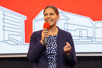 Eine Frau mit hochgesteckten Haaren spricht gestikulierend in ein Mikrofon. Sie trägt ein gepunktetes Kleid und einen Blazer. Der Hintergrund zeigt den Ausschnitt eines Bildschirms.
