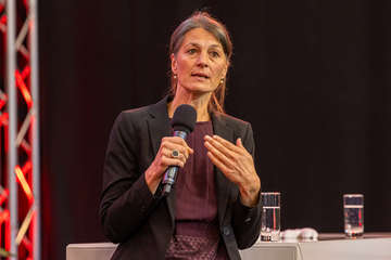 Eine Frau mit hochgebundenen Haaren spricht in ein Mikrofon. Sie trägt ein Kleid und darüber einen Blazer. Sie gestikuliert leicht beim Sprechen.