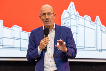 Ein Mann mit Brille und Anzug spricht in ein Mikrofon und gestikuliert dabei. Der Hintergrund des Bildes zeigt einen Ausschnitt eines Bildschirms, auf dem die Ämter in stilisierter Form abgebildet sind.