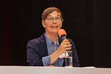 Eine Frau mit Brille und Kurzhaarschnitt spricht in ein Mikrofon und lächelt dabei. Sie trägt eine gestreifte Bluse und einen Blazer.