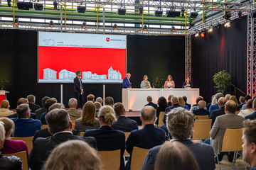 Im Hintergrund des Bildes ist eine Bühne mit großem Bildschirm zu sehen. Vier Personen stehen hinter einem weißen, breiten Stehtisch. Eine Person steht etwas abseits. Im Vordergrund des Bildes sitzt das Publikum, die Blicke sind auf die Bühne gericht