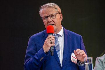 Ein Mann mit kurzen, hellen Haaren und Brille spricht in ein Mikrofon. Er trägt Anzug und Krawatte und gestikuliert leicht.