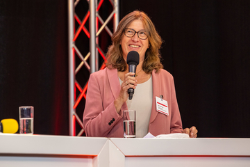Eine Frau mit schulterlangem Haar und Brille steht hinter einem Stehtisch. Sie trägt einen Blazer, vor ihr steht ein Glas Wasser. In der Hand hält sie ein Mikrofon, sie lächelt.