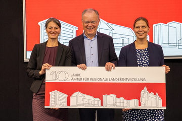 Drei Personen stehen in einer Reihe und lächeln. Sie halten ein Schild, das die Aufschrift "10 Jahre Ämter für regionale Landesentwicklung" trägt und die vier Ämter in stilisierter Form zeigt.