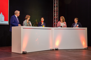 Fünf Personen stehen hinter großen, weißen Stehtischen, die von Bodenstrahlern beleuchtet werden. In der Mitte steht Ministerin Osigus und spricht in ein Mikrofon. Die Landesbeauftragten stehen links und rechts von ihr und schauen sie an.