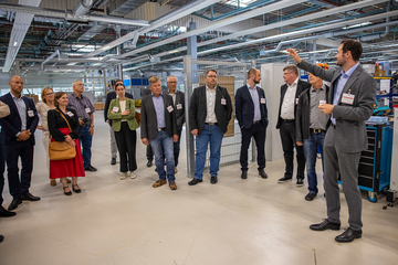 Das Bild zeigt eine große Industriehalle, im Vordergrund steht eine Gruppe von Menschen im Halbkreis. Die meisten Personen schauen zu Christoph Imdahl. Er steht rechts im Bild und deutet auf die Halle.