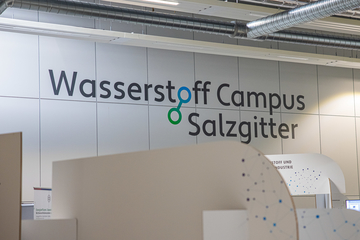 Der Schriftzug Wasserstoff Campus Salzgitter steht in großer Schrift auf einer weißen Wand. Im Vordergrund stehen Pappaufsteller, nur unscharf zu erkennen.