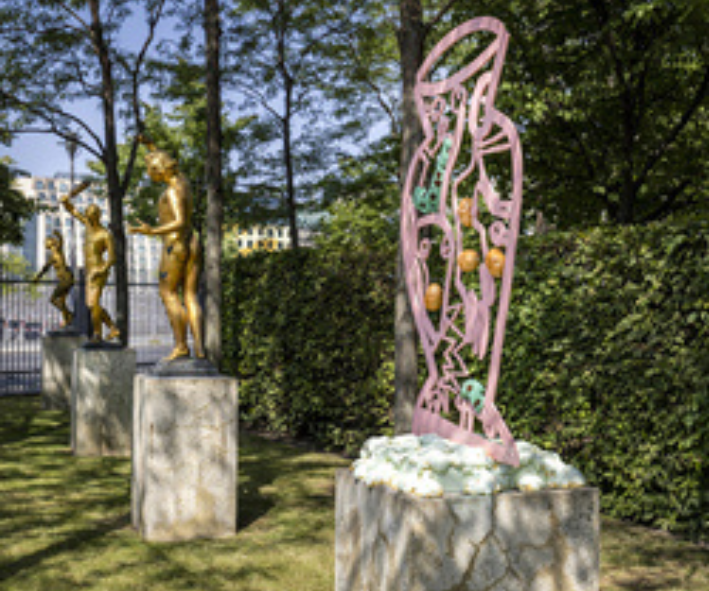 Eine zweidimensionale Skulptur einer antiken Vase mit unterschiedlichen Farben und aus unterschiedlichen Materialien steht auf einem Betonsockel. Im Hintergrund stehen goldene Barockskulpturen, ebenfalls auf Betonsockeln.