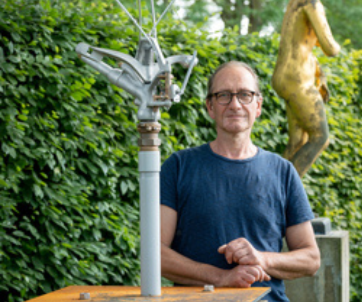 Der Künstler Ingo Schulz steht hinter seiner Skulptur "Trockenübung Solo". Im Hintergrund steht eine vergoldete Barockstatue auf einem Sockel.